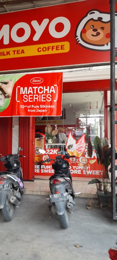 A storefront for "MOYO MILK TEA COFFEE" in Crewek, Indonesia, is captured. The shop's facade features a prominent red sign with the brand name and a cartoon mascot. A banner advertising "MATCHA SERIES" is displayed, detailing "10μm of Pure Silkiness from Japan." In front of the shop, two motorcycles are parked. The one on the left has a license plate reading "10 AFF 05/27". The one on the right has a license plate that reads "K 5241 UP". The environment is outdoors, with a red metal gate to the left of the storefront and a dark metal pole to the right. The ground is paved with concrete.