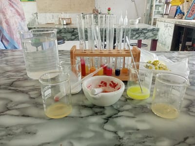 The image depicts a science experiment setup on a marble-patterned countertop. In the center, a wooden test tube rack holds several test tubes containing liquids of various colors (red, orange, yellow, blue). Around the rack are beakers and a mortar and pestle. A large beaker filled with clear liquid stands to the left. A small amount of yellow liquid is in the beaker to the left of the mortar. The mortar and pestle contain a red substance. To the right of the mortar and pestle are two more beakers, one with a yellow liquid and one with a small amount of cloudy liquid. Behind the setup is a lab setting with countertops, stools, and equipment. The location, Puntimatangkuli, Indonesia, is not directly evidenced in the image.