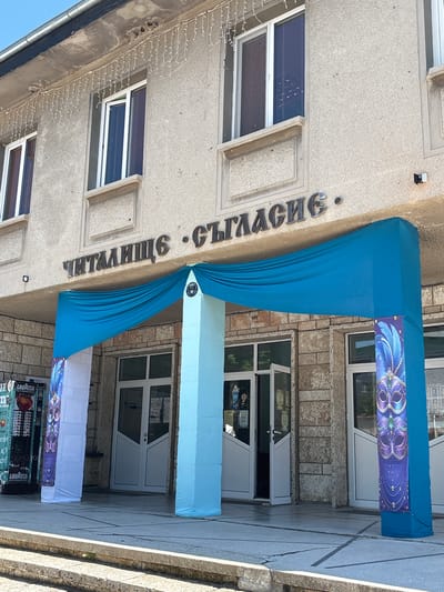The image shows the entrance to a building with a sign in Bulgarian that reads "ЧИТАЛИЩЕ СЪГЛАСИЕ". This translates to "Chitalishte Saglasie", which is a community center or cultural club. The entrance is decorated with draped blue fabric and two tall pillars covered in light blue fabric, with banners on either side featuring purple masks. To the left of the entrance, a vending machine is visible. The building appears to be made of concrete and stone, with several windows on the upper floor. The overall scene suggests a festive or celebratory atmosphere, possibly related to a carnival or masquerade. The location is identified as the Chitalishte Saglasie in Kavarna, Bulgaria.