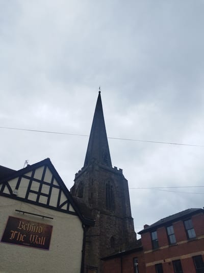 An outdoor photograph depicts architectural structures under an overcast sky in Hereford, United Kingdom. The central and dominant subject is the tall, dark grey stone spire of St Peter's Church, located on St Owen Street, which ascends from a square, crenellated tower featuring arched belfry windows. The spire culminates in a slender point topped with a weathervane. To the left of the church, partially visible, is a building with a white, textured render on its lower section and a dark brown half-timbered gable above, beneath a dark tiled roof where a television antenna is present. A rectangular sign, angled slightly downward, is affixed to the white wall, displaying "Behind The Wall" in ornate dark red lettering and "Freehouse" in smaller script below. To the right of the church, a red brick building with horizontal yellow brick banding and multiple dark-framed windows is partially visible. Two thin, dark utility lines traverse horizontally across the upper middle portion of the frame. The sky is uniformly grey with dense cloud cover. No individuals or discernible activities are present within the scene.