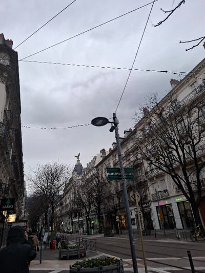 An urban street scene on an overcast day. Tall buildings line the street, interspersed with bare trees. A streetcar line runs along the road, supported by poles with traffic signals. People walk along the sidewalk. A building with an angel statue on top is visible in the distance. The image is rotated counter-clockwise.