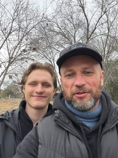 The image depicts a selfie taken outdoors, featuring two individuals. The person in the foreground on the right is a middle-aged man with a beard, wearing a black baseball cap and a dark jacket over a blue sweater. The person on the left is a younger man with wavy hair, smiling and wearing a dark jacket. They are positioned in front of bare trees, suggesting a cool or winter season. The background shows dry grass and some fencing, indicating a rural or semi-rural setting. The overall scene suggests a casual outdoor activity. The image was taken in Comuna Estancia Vieja, Argentina.