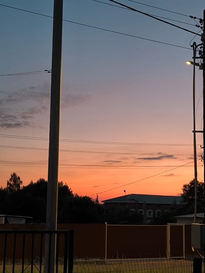 The image depicts a rural scene in Uzunovo, Russia, at dusk. The sky transitions from a deep blue at the top to vibrant shades of orange and pink near the horizon, indicating sunset. A few scattered clouds are visible, tinged with the colors of the setting sun.
Dominating the foreground are utility poles with overhead wires, suggesting the presence of electricity and telecommunication infrastructure. One pole on the left is tall and slender, while another on the right is thicker and carries a street lamp that is illuminated, casting a warm glow.
In the middle ground, a dark silhouette of trees forms a dense line. Behind the trees, a building with a green roof and multiple windows is partially visible. The building appears to be residential, with a distinct architectural style. A brown fence runs horizontally across the lower part of the image, with a metal gate on the left. The ground is covered in grass and some dry vegetation.
The overall mood is peaceful and serene, typical of a quiet evening in the countryside. The warm colors of the sunset create a comforting and picturesque atmosphere. There are no visible people or text in the image. The setting appears to be a residential area on the outskirts of a village or town, characterized by its houses, fences, and utility infrastructure.