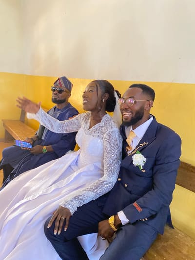 A recently married couple poses for a photograph indoors, alongside a third individual. The groom, positioned slightly above and behind the bride, wears a dark blue suit, eyeglasses, a white flower boutonniere, and a gold watch on his left wrist. His suit jacket has a patch on the sleeve that says "SOSILA DINO". The bride is wearing a white, long-sleeved, beaded wedding gown. To the left, a man wearing sunglasses and a traditional striped cap stands with his right hand outstretched towards the camera, and he is holding a mobile phone in his left hand. The setting appears to be an indoor space, possibly a wedding venue, with a yellow wall. No specific details confirm the location as Kuru, Nigeria.