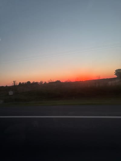 A landscape photograph depicts a vivid sunset. The sky graduates from pale blue to a fiery orange hue on the horizon, behind a line of trees. Power lines stretch across the sky on the left. To the right, the foreground includes a dark asphalt road with a white lane marker, creating a sense of perspective. The image is vertically oriented.