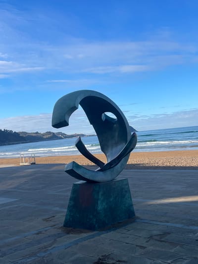 The image displays a large, dark, abstract metal sculpture centrally located on a paved promenade in Zarautz, Spain. The sculpture, with a dark, possibly patinated, metallic finish, exhibits a complex, curved, and open form, featuring internal voids and supporting elements, and rests on a rectangular base of similar material. Immediately behind the promenade is a wide, light-brown sandy beach. Beyond the beach, the blue ocean extends to the horizon, with several white-capped waves breaking towards the shore. Distant, indistinct figures are faintly visible in the water. A white, elevated, multi-tiered structure is present on the sand to the left of the frame. In the far background, rolling green hills line the coastline, with a distant town or village featuring light-colored buildings situated on a headland to the left. The sky is a clear, bright blue with scattered white cumulus clouds. The scene is illuminated by strong sunlight, casting distinct shadows on the promenade surface. No specific individuals or immediate interactions are clearly identifiable in the foreground.
