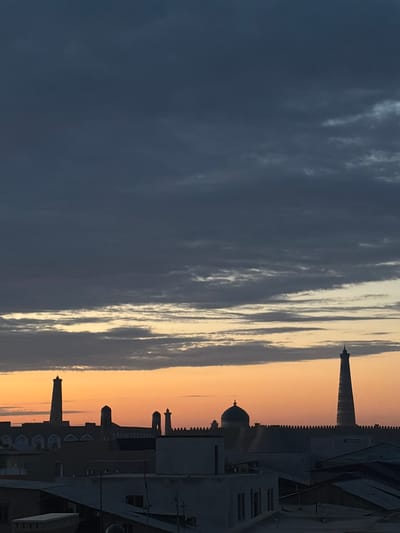 This is a photograph taken from an elevated position overlooking the city of Khiva, Uzbekistan, likely during sunrise or sunset. The sky is a dramatic mix of dark, brooding clouds in the upper portion, transitioning to lighter, wispy clouds illuminated by the warm hues of orange and yellow from the setting or rising sun near the horizon.

Silhouetted against the colorful sky are the iconic architectural features of Khiva, a UNESCO World Heritage site. Dominating the skyline are several minarets and domes, characteristic of Islamic architecture. Most prominent is a tall, slender minaret on the right, tapering towards its peak, possibly the Islam Khoja Minaret. To its left, a dome with a finial is visible, likely part of a mosque or mausoleum. The city itself appears to be comprised of low-rise buildings, many with flat roofs and traditional architectural details, all rendered in silhouette. A fortified wall, characteristic of the ancient city, runs horizontally across the mid-ground, behind which the minarets and domes stand. The foreground shows the rooftops of more recent structures, some appearing to be simple, modern buildings with flat or angled roofs, partially obscuring the view of the ancient city. There are no discernible people or animals in the frame. The overall impression is one of ancient history meeting the present, set against a striking sky.