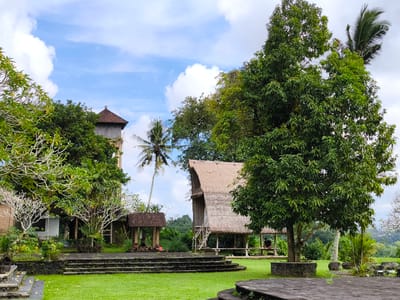 The image depicts a tranquil scene in Sidemen, Indonesia. Lush green grass covers the foreground, leading to a series of stone steps. A traditional thatched-roof hut with a raised platform and wooden support beams sits amidst the greenery, with a person seated inside. Nearby is a smaller, open-sided pavilion with a tiled roof. A mix of tropical trees and vegetation surrounds the structures, including a tall palm tree. The sky is light blue with scattered white clouds. A multi-story building with a brown roof is partially visible behind the trees.
