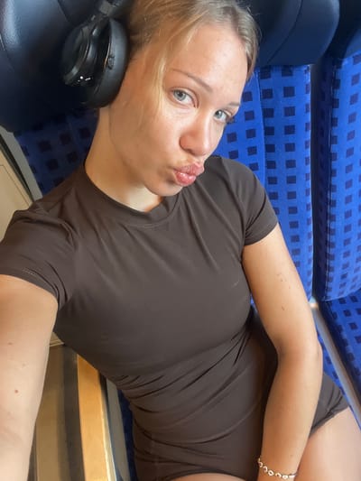 A young blonde woman with blue eyes is seated on a blue upholstered seat, possibly on a train, given the pattern of the upholstery. She is wearing a dark brown short-sleeved top and appears to be wearing matching shorts. On her head, she has large black headphones. Her lips are puckered, suggesting she is making a kissing face. She is looking towards the camera. A beaded bracelet with small flower-shaped charms is visible on her right wrist. The background consists of the seat she is sitting on and the seats behind her, which are blue with a dark blue checkered pattern. The lighting suggests it is daytime. The image does not provide specific location details for Ampfing, Germany; it appears to be a selfie taken on public transportation.