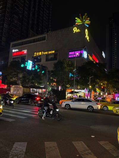 The image captures a vibrant night scene in an urban area, likely a commercial district in 天宫殿街道, China. The perspective is from the side of a road, looking towards a multi-story building illuminated by numerous neon signs.
The street is bustling with activity, featuring several vehicles including a white car, a black car, and at least two yellow taxis, one parked and one further down the road. Multiple motorbikes or scooters are visible, with riders waiting on the street, suggesting traffic or a stop at a crosswalk. A pedestrian crosswalk with white stripes is prominent in the foreground. Trees with decorative star-shaped lights line the street, adding to the festive nighttime ambiance. Traffic lights displaying green signals can be seen.
The prominent building on the left is adorned with various bright signs. At its peak, a large yellow sign reads "9st." (likely "9th st.") accompanied by a colorful, pinwheel-like sculpture. Below this, a neon sign displays "HICKEY" alongside Chinese characters. Further down, another sign reads "空瓶子 LIVE SHOW" (Empty Bottle LIVE SHOW), suggesting an entertainment venue. Other visible text includes "无界台" (Wujie Tai - Boundless Stage) and "HOOJA" with "合家" (He Jia - Family). A tall, darker building stands in the background to the left. The scene is vividly lit by artificial lights, creating a dynamic night atmosphere.