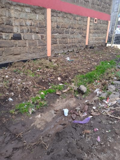 The scene depicts an outdoor environment in Njoro ward, Kenya. The foreground and midground consist of disturbed dark earth mixed with dry vegetation, sparse green grass, small stones, and various forms of litter. Observable debris includes crumpled plastic bags (one light blue/white, one purple net-like), white plastic fragments, paper scraps, and unidentifiable refuse. A stone or brick wall, composed of dark, irregularly shaped blocks, forms the background. The wall features a horizontal red painted stripe across its upper section and two complete, vertically oriented orange painted stripes, along with a partial third stripe on the right. A small, square, orange-painted recess is present on the upper right portion of the wall. Beyond the wall's right edge, a utility pole and a partially visible sign with text including "KIN" and "Welcome" are discernible. No individuals, explicit actions, or interactions are visible.