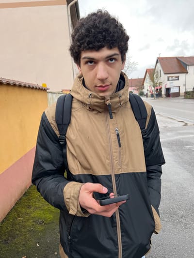 A young male individual, appearing to be in his late teens or early twenties, with dark, curly hair and light-to-medium complexion, is depicted in a medium-shot portrait taken outdoors in Meistratzheim, France, under an overcast sky. He is wearing a two-tone jacket, light tan/brown on the upper portion and black on the lower half, which exhibits dark specks or stains on the tan fabric. A black backpack is worn over his shoulders. His right hand holds a black smartphone horizontally, with the screen oriented away from the camera. He gazes directly forward with a neutral to slightly serious expression.
The immediate foreground to the left features a section of a building with an orange-tan painted wall and a terracotta-tiled roofline. Green moss is visible on the ground level adjacent to this structure. To the right and extending into the background, a paved road surface is present. Beyond the road, architectural elements characteristic of the Alsace region are visible. The most prominent building on the right has a brown tiled roof, light-colored walls, and appears to be a commercial establishment, possibly an inn or restaurant, with a sign partially displaying "Gaststätte." Another building with a red tiled roof is visible further in the background. Sparse, bare trees are discernible in the distance, and the sky is uniformly grey.