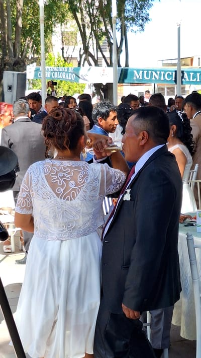 The image depicts a wedding celebration occurring outdoors in Cerro Colorado, Peru. The bride and groom are prominent, with the groom in a dark suit and the bride in a white dress. They are drinking from a champagne glass. A crowd of people, including family and guests, surround the couple. A banner with "MUNICIPALIDAD DE CERRO COLORADO" is visible. Trees are present, suggesting a park setting.