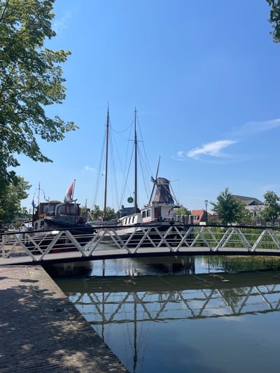 A wide shot captures a canal in Meppel, Netherlands, on a sunny day with a clear blue sky. A white pedestrian bridge with a geometric railing spans the water in the foreground, reflecting on the calm surface. Beyond the bridge, several boats are docked, including a larger sailboat with tall masts and a motorboat. In the background, a traditional Dutch windmill stands behind the boats, adding to the picturesque scene. Buildings with red-tiled roofs and trees line the canal bank. A large tree with green leaves shades the left side of the image. The scene is tranquil and scenic, showcasing a characteristic Dutch waterway.