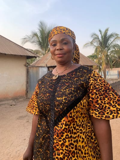 An adult Black woman wearing a leopard print headscarf and dress stands outside. Her dress is leopard print with a black lace overlay on the front. She is also wearing a gold necklace and earrings. A corrugated metal roof, a concrete wall, and a palm tree are visible behind her. The sky is light blue and the ground is tan. The photo is slightly tilted to the left.
