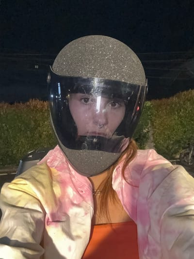 A woman in a tie-dye jacket and orange top is wearing a silver glitter motorcycle helmet with a clear visor. Her face is visible through the visor, and she appears to have a nose ring. The background includes a dark, possibly nighttime, scene with vegetation and what may be a street. The location is Kerobokan Kelod, Indonesia.