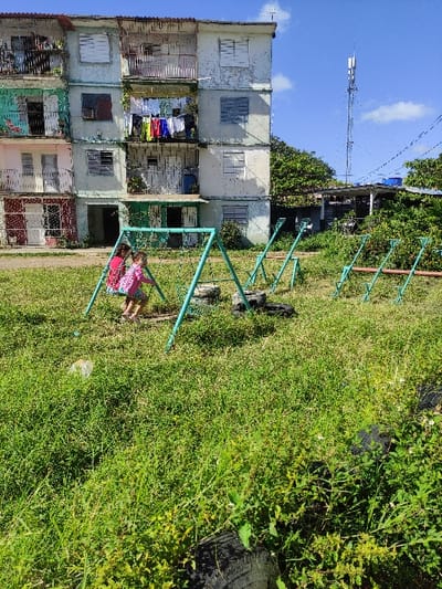 The image depicts an outdoor scene in Ciego de Ávila, Cuba, under a clear blue sky with sparse clouds. In the foreground and midground, a patch of unkempt green grass and weeds covers the ground. Two young children are present; one child, wearing a red shirt, sits on a teal-colored metal swing, while another child, in a pink shirt, stands beside the swing, appearing to interact with it.
The children are situated on a dilapidated playground area featuring the operational swing set and several other teal-painted metal frames of what appear to be disassembled or partially buried playground equipment. Multiple black, tire-like objects are embedded or scattered within the overgrown grass.
In the background, a weathered multi-story apartment building rises, displaying a mix of faded white, light blue, pink, and green paint on its facades. Several windows are visible, many equipped with closed louvered shutters. Balconies protrude from various levels; on one balcony, colorful laundry, including red, blue, and white garments, hangs to dry. A corrugated metal awning covers a ground-level entrance.
To the right of the apartment building, a dense cluster of trees partially obscures a smaller structure with a metal roof. Beyond these trees and the building, a tall, metal lattice telecommunications tower with multiple antennas stands prominently against the sky. Utility lines are visible traversing the upper right quadrant of the image. A blue water tank on a platform is also visible in the background, further to the right.