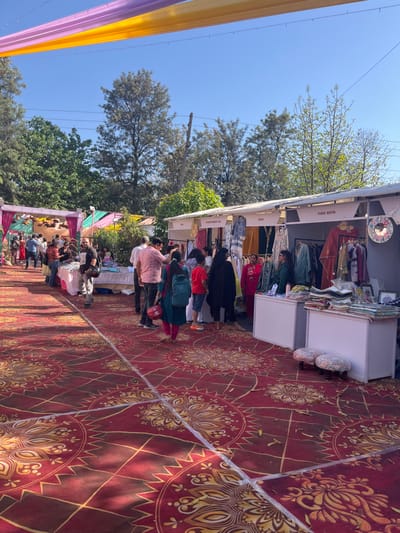 The scene takes place in a marketplace in Sector 17, Chandigarh, India, located at latitude: 30.764, longitude: 76.798. The image depicts a bustling marketplace with several stalls, each showcasing a variety of clothing items. There are numerous individuals, both men and women, browsing the displayed merchandise. Some are engaged in conversations, likely discussing the clothing or negotiating prices. The marketplace is adorned with colorful canopies, likely for shade and to enhance the overall aesthetic. The ground is covered with a red patterned carpet, adding to the vibrant ambiance. The vendors are dressed casually, reflecting the informal atmosphere of the market. Several small, white, square stools are placed in front of the stalls.  The stalls themselves are well-stocked with clothing items, neatly displayed on racks.  The image captures a moment of lively interaction between the vendors and customers, highlighting the vibrant nature of this particular market.