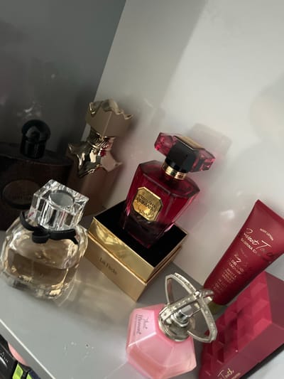 The image displays a collection of perfume bottles and a body lotion tube arranged on a shelf. The perfumes are of various shapes and colors, including a dark brown bottle with a black cap, a gold-capped bottle with an ornate design, a clear bottle with a faceted crystal cap, a deep red bottle with a faceted stopper, and a light pink faceted bottle with a decorative silver atomizer. A red tube of body lotion with white text is positioned to the right of the red perfume bottle. A dark red, faceted object, possibly another perfume bottle or case, is visible in the lower right corner. The shelf appears to be made of a grey material. The background consists of a light grey and a darker grey surface, suggesting an indoor setting. There are no people or visible actions in the image. The scene is static and focuses on the arrangement of the cosmetic products. The location of Ognyanovo, Bulgaria is not depicted in this image.