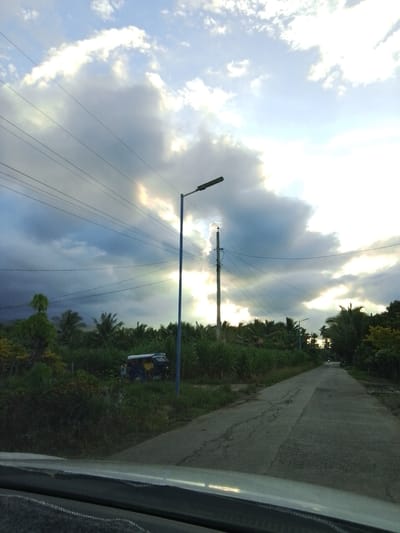 The image depicts a rural road in Santa Cruz, Philippines, under a dramatic sky filled with dark, billowy clouds interspersed with patches of bright sunlight. Several power lines crisscross the scene, supported by utility poles. The road itself is paved but shows signs of wear and tear, with cracks and uneven surfaces.
To the left of the road, dense vegetation including tall grasses and palm trees lines the area. A blue and yellow trishaw, a common mode of transport in the Philippines, is parked on the side of the road amidst the greenery. A tall, blue lamppost stands prominently on the left side of the road.
Further down the road, more trees and vegetation are visible, with a few houses and structures partially obscured by the foliage. The road curves gently into the distance, where the sky appears brighter. The overall impression is one of a tropical setting at dusk or dawn, with the weather contributing to a slightly moody atmosphere. The perspective is from a vehicle, indicated by the dashboard and windshield visible in the foreground. There are no people clearly visible in the image.