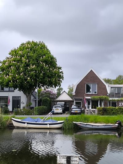 The photograph depicts a canal scene in Oudorp, Netherlands, under an overcast sky. Two boats are moored along the canal bank. The first boat, positioned in the left foreground, is white with a navy blue trim and a blue cover. The second boat, on the right, is gray with an outboard motor attached at the stern. Both are reflected in the water.
Behind the boats, the scene transitions to the land with manicured grass and residential buildings. A large tree with white blossoms dominates the left side of the frame, partially obscuring a light-colored house labeled with the number "26." To the right of the tree, a lilac bush adds a touch of purple to the green landscape. A white car and a dark car are parked in the driveway.
To the right of the cars, a brick house with a steep gable roof is visible, labeled with the number "27." The house has a prominent square window on the second story and greenery surrounding the lower level. A wooden gate is partially visible in front of the house. A Netherlands flag is flown from the front of the property.
In the water in the foreground, a partially submerged signpost is visible, displaying writing, including the word "SOLEIL" and the license plate "42-12-YT."
