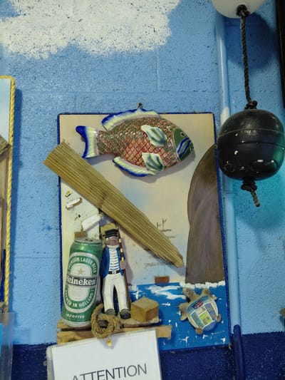 The image shows a nautical-themed display affixed to a textured blue wall. A black buoy hangs from the top. The display features a ceramic fish, a wooden plank, a miniature turtle figurine, a toy sailor, and a Heineken beer can. An "ATTENTION" sign is also present. It is likely a decorative display in Cutler Bay, Florida.