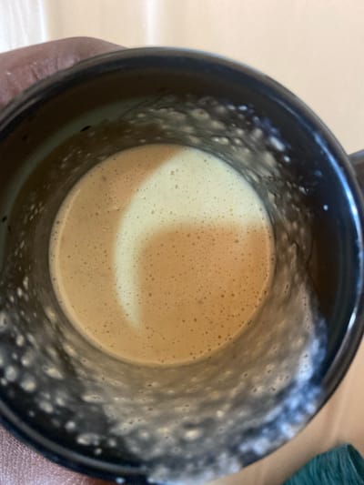 A close-up, top-down shot captures the interior of a dark-colored, possibly black, ceramic mug. The mug contains a light brown, creamy beverage, likely a coffee or tea drink with foamed milk or creamer. The surface of the liquid is covered in a dense layer of small, uniform bubbles, forming a rich foam. Within this foam, a distinct, lighter-toned, crescent or C-shaped pattern is visible, suggesting a deliberate design or a natural swirl. The inside walls of the mug show scattered residue or foam adhering to the surface, indicating recent use or agitation of the liquid. The background is out of focus, revealing a light-colored surface, possibly a tabletop, and a small, blurred section of a green or teal object to the right. The image depicts a prepared beverage in Lakka, Sierra Leone. No text is visible.