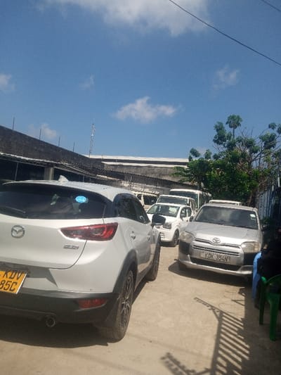The scene depicts an outdoor vehicle parking area in Majengo ward, Kenya, under a clear blue sky with scattered white cumulus clouds. A white Mazda CX-3 crossover SUV, license plate KDV 85C, is prominently parked on the left, viewed from its rear-left quarter. To its right and slightly forward is a silver Toyota Probox van, license plate KDK 354N, facing the viewer. Multiple other white vehicles, including a small hatchback and various passenger vans, are parked in rows extending into the background. On the far right, a person is partially visible, seated on a green plastic chair, wearing dark clothing. The ground is paved with light-colored concrete or asphalt, displaying distinct shadows cast from the right, including a patterned shadow consistent with a fence or railing. In the background, a building with a dark, sloping roof is on the left, behind which another light-colored building and a tall metal communication tower are visible. Green trees and foliage are present on the right, partially obscuring a blue corrugated metal fence.