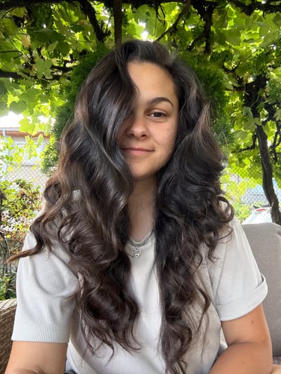 A young woman with long, wavy dark brown hair is captured in a medium shot, seated outdoors. Her hair is styled with some strands falling over her face. She is wearing a light-colored, ribbed short-sleeved shirt and a necklace with a heart-shaped pendant. She is positioned in front of a backdrop of lush green foliage, suggesting a natural or garden setting. Part of a woven chair and a couch cushion are visible in the foreground and to the right, respectively. The background also includes a chain-link fence and some buildings, indicating an urban or suburban environment. The image appears to have been taken in Oreshak, Bulgaria, based on the provided context.