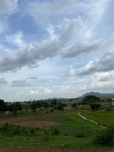 An overcast sky with dramatic, textured clouds dominates the upper portion of the image. Below, a panoramic view of a rural landscape unfolds, featuring rolling hills, scattered trees, and cultivated fields. In the mid-ground, a narrow, flowing stream cuts through a vibrant green field, with a lone figure walking alongside it. A few small structures, possibly homes, are visible in the distance, nestled amongst the trees. The overall impression is one of a serene, agricultural setting in Rimin Ziem, Nigeria, under a dynamic sky.