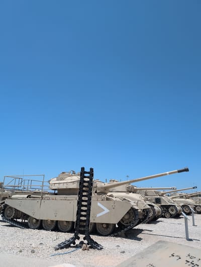 A clear, bright day under a brilliant blue sky. The scene is dominated by a line of tan-colored armored vehicles, appearing to be tanks, parked in an outdoor lot. The ground is covered with a light-colored gravel. The foremost vehicle, larger than the others, has a long cannon pointing towards the right side of the frame. A black, ladder-like object stands in front of this tank, with a white chevron marking visible on the tank's side. Further back, more tanks are visible in a procession, their cannons also extended. The environment suggests a military museum or outdoor exhibition, with a clear sky and open space. The image is taken from a low angle, emphasizing the imposing size of the vehicles.