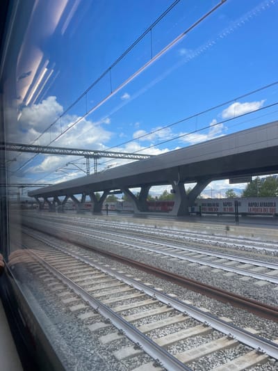The image depicts an exterior view of the Napoli Afragola high-speed train station in Afragola, Italy, observed from inside a train. Multiple railway tracks with light grey ballast and concrete sleepers occupy the foreground and midground, extending towards the horizon. Above the tracks, a complex system of overhead catenary lines and supporting gantries is visible.

In the midground, the station platform features a long, modern grey canopy supported by distinctively angled, geometric pillars. Large white banners with black text are visible on the platform, including "NUOVA LINEA AV/AG", "NAPOLI-BARI IN 2 ORE", and "NAPOLI AFRAGOLA LA PORTA DEL SUD". No individuals are present on the platform or tracks.

The upper portion of the image shows a bright blue sky with scattered white cumulus clouds. Reflections of interior elements, possibly overhead lighting, from the train cabin are visible in the top-left quadrant of the window. Distant green foliage and hazy mountains are discernible beyond the station structure. The scene is illuminated by natural daylight.