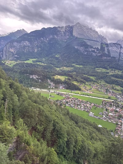 The image depicts a panoramic view of a mountainous region in or around Hüsen, Switzerland, on an overcast day. In the foreground, a steep, densely forested mountainside descends sharply. The trees are predominantly evergreen, with varied shades of green. The middle ground reveals a valley with a small village, characterized by numerous residential buildings with red-tiled roofs, interspersed with green fields and patches of forest. A road or pathway is visible, weaving through the village and fields. In the background, imposing, rugged mountains with rocky outcrops and some snow-capped peaks rise sharply against the cloudy sky. The overall impression is one of a remote, picturesque alpine landscape with a human settlement nestled within it. There are no people or animals visible in the frame.
