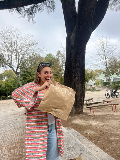 A young adult female, approximately 20-30 years old with light brown hair and sunglasses resting on her head, stands in an outdoor park setting. She wears a long, open cardigan with horizontal stripes in white, pink, orange, green, and grey, over a white top and light blue denim jeans. Her expression is an open-mouthed smile, appearing joyful. She holds a brown paper shopping bag branded "PARFOIS" with "PARFOIS.COM" at the bottom, open at chest height.
The environment features a large tree with a bifurcated trunk on the right, and scattered trees, some deciduous with bare branches and others with green foliage, under an overcast sky. A paved area is visible on the left, transitioning to a dirt ground on the right. In the mid-ground, multiple rustic wooden benches with red legs are present, along with two parked bicycles. In the background, a distinctive modern building with a multi-faceted light green roof structure, identifiable as the Casa da Música, is visible on a raised platform. This location is within the park surrounding the Casa da Música, situated in the Boavista area of Lordelo do Ouro e Massarelos, Porto, Portugal.