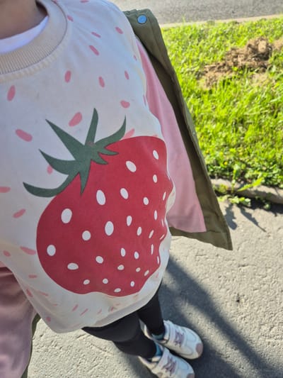 The image shows a close-up view of a person wearing a light-colored sweatshirt with a large red strawberry graphic and scattered pink dots. A green jacket is partially visible over the sweatshirt, and the person is wearing black leggings and white sneakers with pink laces. The scene appears to be outdoors, on a paved surface, with green grass and some dirt visible in the background. The lighting suggests it is daytime, and shadows are cast on the ground. The specific location in Petrozavodsk, Russia cannot be determined from the visual information provided.