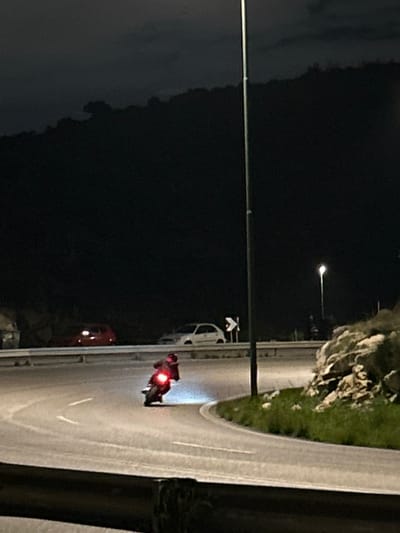 The image depicts a nighttime scene on a curving road in Anatoliki Attiki, Greece. The central subject is a person riding a motorcycle on the right side of the asphalt road, moving away from the viewer. The motorcycle's bright red taillight is distinctly illuminated, and the rider is wearing a helmet and appears to have a large item, possibly a delivery box or backpack, on their back.

The road curves gently towards the right, with white lane markings visible on the asphalt. To the right of the road, a rocky hillside covered with some grass is present, separated by a guardrail. A tall, slender streetlight pole stands on the left side of the road. Another illuminated streetlight is visible further in the background, casting light on parts of the road and the hillside.

In the background, other vehicles are present. A white car is parked off to the right side of the road, further ahead of the motorcycle. Near the white car, a dark road sign with a white chevron pointing left is visible. Further down the road on the left, a red car can be faintly seen. The sky is dark, indicating night, and the artificial lighting creates a moody, quiet atmosphere. No discernible text is visible in the image.