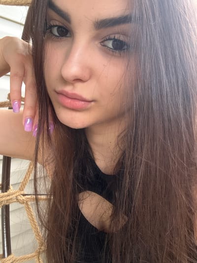 The media file is a close-up, low-angle, portrait-style photograph of a young woman. She has long, dark brown hair that frames her face and falls across the frame. Her face is turned to the side, with her left eye prominently visible and her right eye partially obscured by her hair. She has dark eyebrows, long eyelashes, and her skin appears fair with a subtle sheen. Her lips are slightly parted and appear to be colored with a light pink or nude lipstick. Her right hand is raised and resting near her face, with her fingers gently touching her cheek. Her fingernails are visible and painted with a gradient of purple and pink, suggesting a cosmetic detail. The background is out of focus, but appears to be a dimly lit indoor setting with elements of rope and a light-colored material, possibly indicating she is sitting in a hammock or a chair with rope accents. The overall mood of the photograph is intimate and serene, with the soft lighting and her relaxed pose contributing to this feeling. No text is visible in the image. The context provided indicates the location is Polena, Bulgaria.