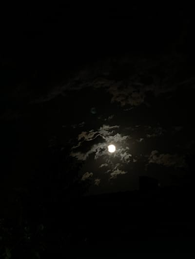 A nighttime scene shows a bright full moon partially obscured by scattered clouds. The surrounding environment is dark with silhouettes of trees and possibly a building to the right. The image appears to be taken outdoors. There is no indication of the location being Koniovets, Bulgaria.