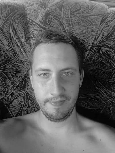 A black and white, close-up, selfie-style portrait of a bare-chested Caucasian male lying on a patterned sofa in Vil'nokur'yanivs'ke, Ukraine. The man has light brown hair, blue eyes, and a short beard. He is looking directly at the camera with a neutral expression. The sofa has a dark, leaf-like pattern.