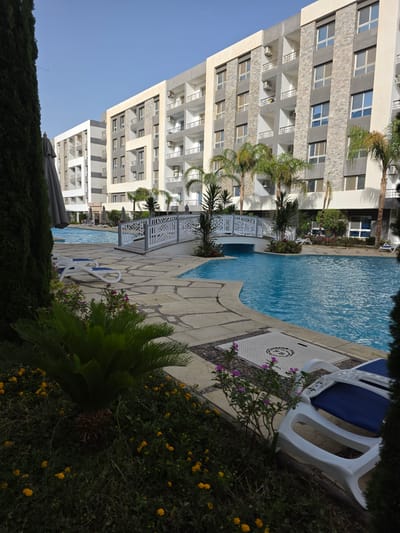 This outdoor scene in Al Ahia', Egypt, features a multi-story building on the left side of the frame, constructed with a gray and white facade. Multiple windows with balconies are visible. Adjacent to the building is a swimming pool filled with blue water. A paved area surrounds the pool. White lounge chairs are positioned next to the pool. Lush green plants and shrubs are in the foreground and surrounding the area, with yellow and purple flowers visible. The sky is a clear blue.