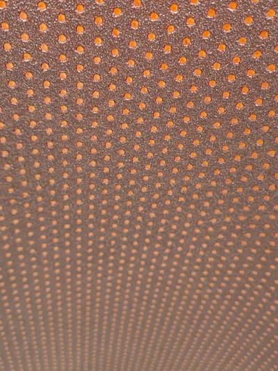 The image shows a close-up, abstract view of a textured surface with an orange-dot pattern. The surface appears to be dark brown or black and is covered in evenly spaced, raised orange dots. The dots vary slightly in size and intensity, suggesting a natural or textured material rather than a perfect print. The lighting creates subtle shadows and highlights on the dots, giving them a three-dimensional appearance. The pattern recedes into the background, creating a sense of depth. There are no people, animals, or recognizable objects in the scene. The image is purely a study of texture and pattern. Without additional context, it is impossible to determine the location of this surface within the city of Settimo Torinese, Italy.