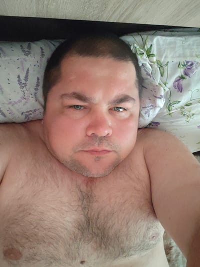 The image is a close-up, low-angle selfie of a bare-chested man lying in bed. He is facing the camera, with his head resting on a pillow. The pillow is white with a floral pattern featuring purple flowers and green leaves. Behind the pillow is a dark headboard. The man has short, dark hair, light green eyes, and stubble on his chin. His expression is neutral. The background consists of a light-colored, textured surface that appears to be a wall or headboard. The lighting suggests it is daytime, likely indoors. The man is in a relaxed position, lying down. There is no visible text in the image. The location context is Русаковское сельское поселение, Ukraine.