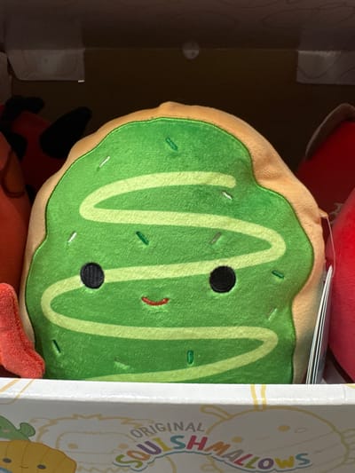 A plush toy resembling a green iced cookie with a smiling face and sprinkles is centrally positioned in the foreground. The toy is light brown around the edges, simulating the cookie dough. It has black button eyes and a small red curved mouth. The green frosting is decorated with a white zig-zag pattern and various colored sprinkles. The plush toy is inside a box, with the lid open, and other plush toys, one red and one orange, are partially visible on either side. The box has "ORIGINAL SQUISHMALLOWS" printed on it. The image is taken from a slightly elevated angle, looking down at the toy. The lighting is dim, suggesting an indoor setting, possibly a store shelf. No specific location in Keila, Estonia can be determined from the image.