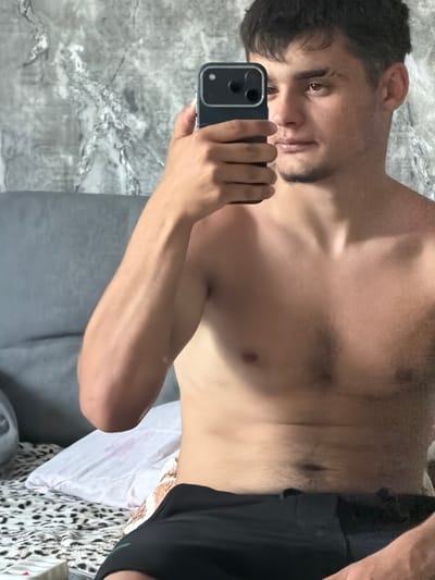 A young man, with short dark hair and a light beard, is taking a selfie in a room. He is shirtless, wearing black shorts. He is holding a dark smartphone in his right hand, pointed towards a mirror. The background shows a textured wall and a grey headboard of a bed, covered with a patterned bedspread and a white pillow. There is a hint of a window or another light source to the left, partially obscured by a pillow. The overall lighting suggests it is daytime. The location is identified as Avdarma, Moldova, though no specific landmarks or city details are visible in the image itself.