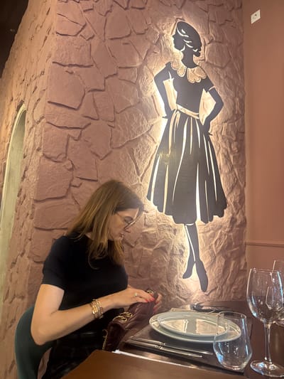 The image depicts an indoor scene, likely a restaurant or dining area, in Torres Vedras, Portugal, illuminated by warm, artificial light, suggesting an evening setting.
The main subject is a woman, positioned on the right side of the frame, seen from the waist up in profile. She has long, light brown hair and wears glasses, a dark short-sleeved top, and dark-colored bottoms. She is looking down into a brown handbag with a gold chain strap, which rests on a table beside her. Her right hand, adorned with a ring, holds the bag, while her left wrist displays multiple golden bracelets.
A large, distinctive black metal silhouette of a woman is mounted on the wall behind her. The silhouette, featuring a woman in a vintage-style dress with a ruffled collar and full skirt, holding what appears to be a tray or dish, is backlit, casting a glow around its edges. The wall itself is textured with a reddish-brown, stone-like pattern.
On the table to the woman's right, a place setting is visible, including two clear wine glasses and two light-colored plates, stacked with a fork and knife. The table surface is dark. The ceiling above is a plain, light beige, with a white electrical outlet seen on it. The overall ambiance appears cozy and decorative. There is no visible text in the image.