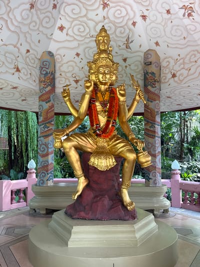 A golden statue of a multi-armed deity is the central element of the image. The statue is adorned with a garland of orange and yellow flowers and is positioned on a maroon base. The deity has multiple faces and is holding various objects in its hands, including a knife and a pot. The statue is situated inside a structure with a ceiling decorated with cloud patterns and floral motifs. Pillars painted with dragon designs support the structure. Beyond the pillars, lush green foliage is visible. The scene appears to be located in Samrong Nuea Subdistrict Municipality, Thailand, based on visual cues associated with Thai religious iconography and architecture.