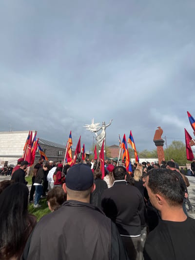 The image captures a gathering of people in Akhuryan, Armenia, likely for a commemorative event or demonstration. Prominently featured are numerous Armenian flags being held aloft, interspersed among the crowd. In the background, two significant monuments are visible: the statue of Mother Armenia, depicted as a silver-colored figure with arms raised, and a darker, blockier sculpture of a bird or other national symbol on a plinth. The sky is overcast, suggesting a cloudy day. People are facing towards the monuments.