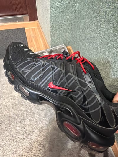 A black and grey Nike Air Max Plus TN sneaker with red laces and a red Nike swoosh logo is held up in front of a carpeted floor, a wooden door frame, and a greenish-grey wall. The location in the city of Kovel, Ukraine is not discernible from the provided image.