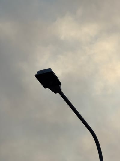 The image shows a black lamppost against a cloudy sky. The lamppost is silhouetted, with its curved pole extending from the bottom right corner upwards towards the top left. The head of the lamppost is a dark, rectangular fixture. The sky is overcast with varying shades of gray and hints of lighter clouds, suggesting either dawn, dusk, or a generally cloudy day. There are no people or other objects visible in the frame. The scene is devoid of any contextual information that would place it in Helsingborg, Sweden, or any other specific city.
