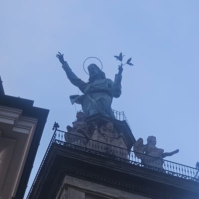 The image captures a statue of Jesus Christ on top of a building in Calcinate, Italy. The statue depicts Jesus with his arms raised and holding a sprig with leaves in his right hand. He is wearing a halo. Around the base of the main statue, there are smaller sculptures of angels. The statue is made of a dark material, likely bronze or stone, and appears weathered. The building itself has decorative architectural elements, including a wrought-iron railing surrounding the base of the statue. The sky behind the statue is a hazy, light blue, suggesting an overcast day or early morning/late evening light. The bottom left of the image shows a portion of an adjacent building with a neutral-colored facade and dark vertical accents.